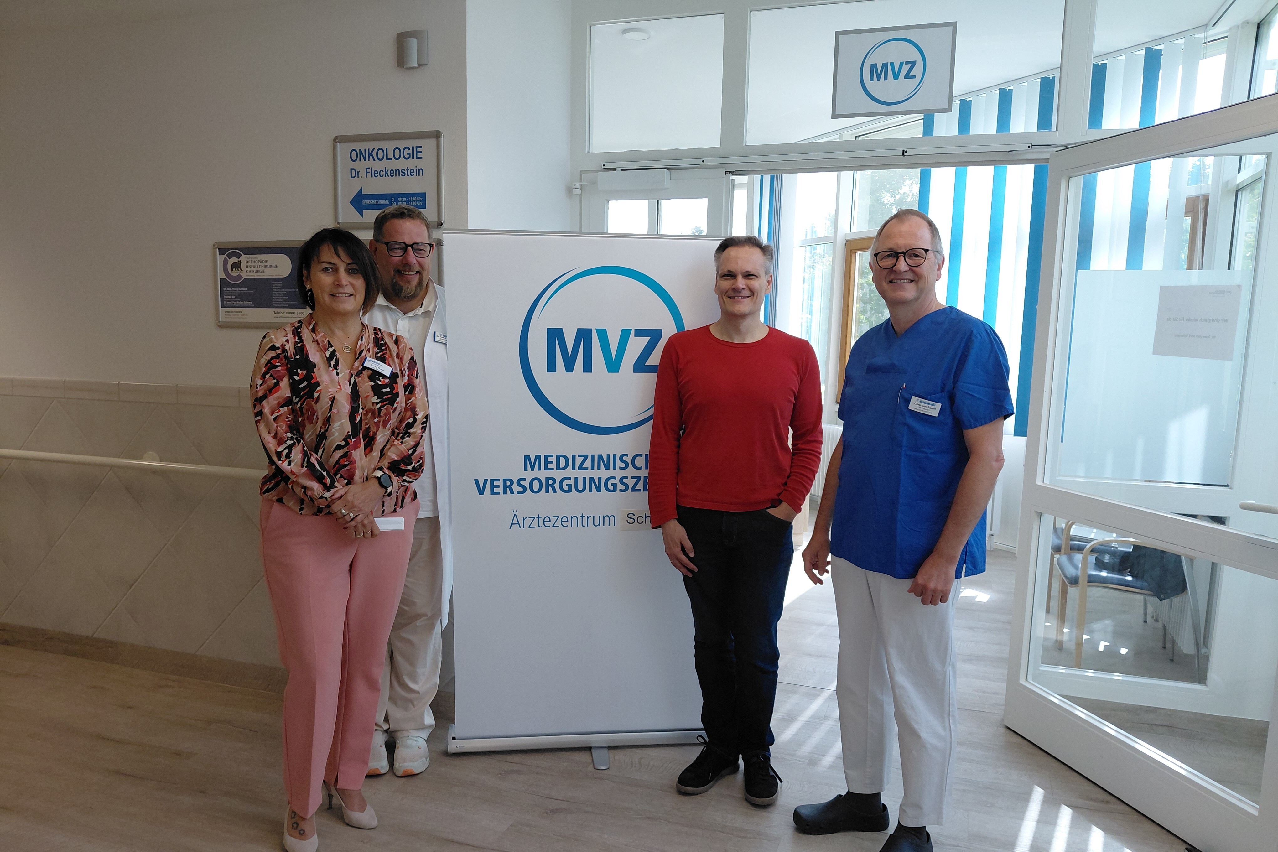 Das Foto zeigt vier Personen, die in einem hellen Flur vor einem Aufsteller mit dem Logo „MVZ – Medizinisches Versorgungszentrum – Ärztezentrum Schongau“ stehen.  Von links nach rechts:  Eine Frau mit dunklen Haaren, die eine rosa gemusterte Bluse und eine hellrosa Hose trägt. Sie lächelt und hat ihre Hände locker vor dem Körper verschränkt.  Ein Mann in weißem Arztkittel steht etwas im Hintergrund, schaut freundlich in die Kamera und trägt eine Brille.  Ein Mann mit kurzem, grauem Haar, der ein rotes Langarmshirt und schwarze Hose trägt, steht in der Mitte. Auch er lächelt.  Ein weiterer Mann, rechts im Bild, trägt eine blaue Kasack-Oberbekleidung und eine weiße Hose – typische medizinische Berufskleidung. Auch er trägt eine Brille und lächelt.  Im Hintergrund ist ein heller Eingangsbereich mit automatischen Glastüren zu sehen. Über dem Eingang hängt ein weiteres MVZ-Schild. Links an der Wand zeigt ein Wegweiser zur Onkologie (Dr. Fleckenstein).