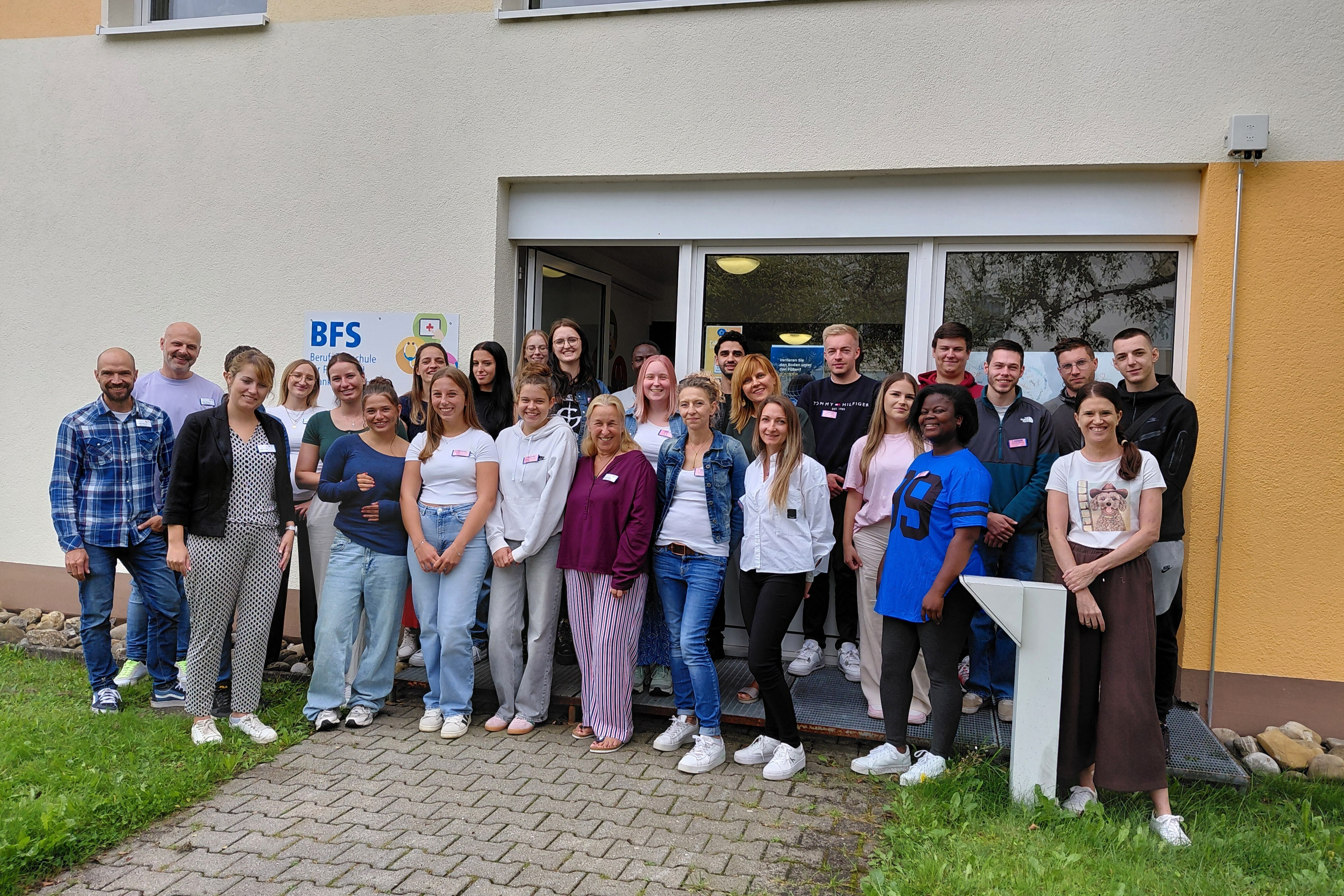 Vor einem Gebäude mit der Aufschrift BFS steht eine größere Gruppe junger Menschen dicht nebeneinander. Es handelt sich überwiegend um Frauen und einige Männer. Die Gruppe trägt legere Kleidung, viele in Jeans, T-Shirts oder Sweatshirts. Alle blicken in Richtung Kamera, einige lächeln. Im Hintergrund ist der Eingangsbereich des Gebäudes mit Glastüren sichtbar. Der Vordergrund zeigt einen gepflasterten Weg und etwas Rasen.