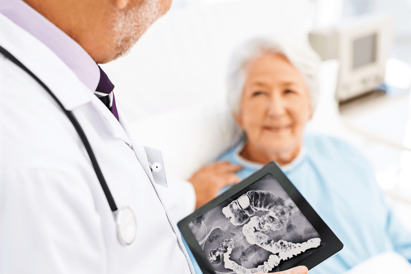Ein Arzt zeigt einer älteren Patientin auf einem Tablet ein Röntgenbild mit Darmkrebs. Die Frau liegt im Bett und schaut den Arzt ernst an.