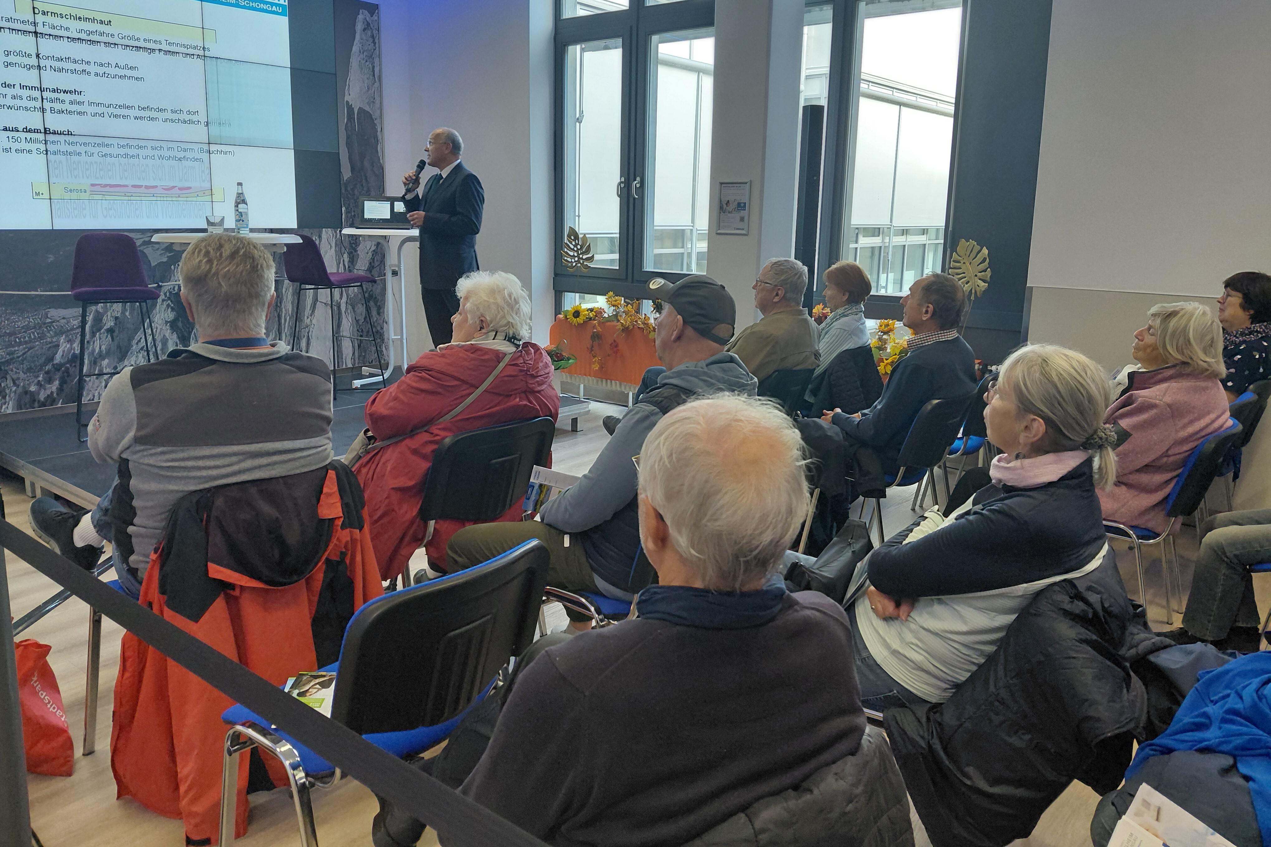 Personen sitzen in einem Veranstaltungsraum und hören einem Vortragenden zu.