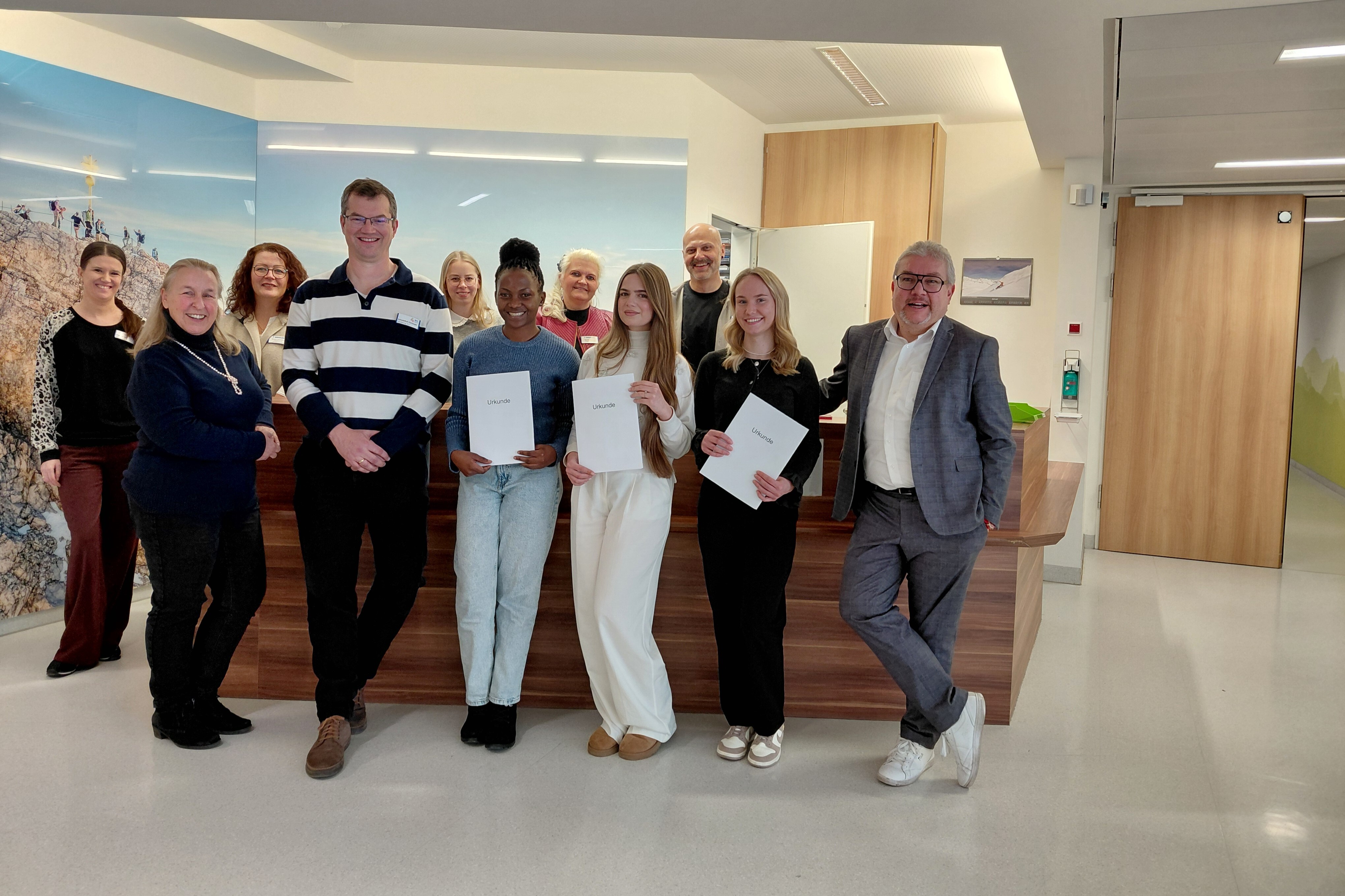 Drei Staatspreisträgerinnen der Pflegeschule stehen in einem Innenraum und halten ihre Urkunden in den Händen. Neben ihnen stehen der Geschäftsführer, die Pflegedienstleiterin, der Schulleiter sowie weitere Vertreter der Berufsfachschule und der Krankenhaus GmbH