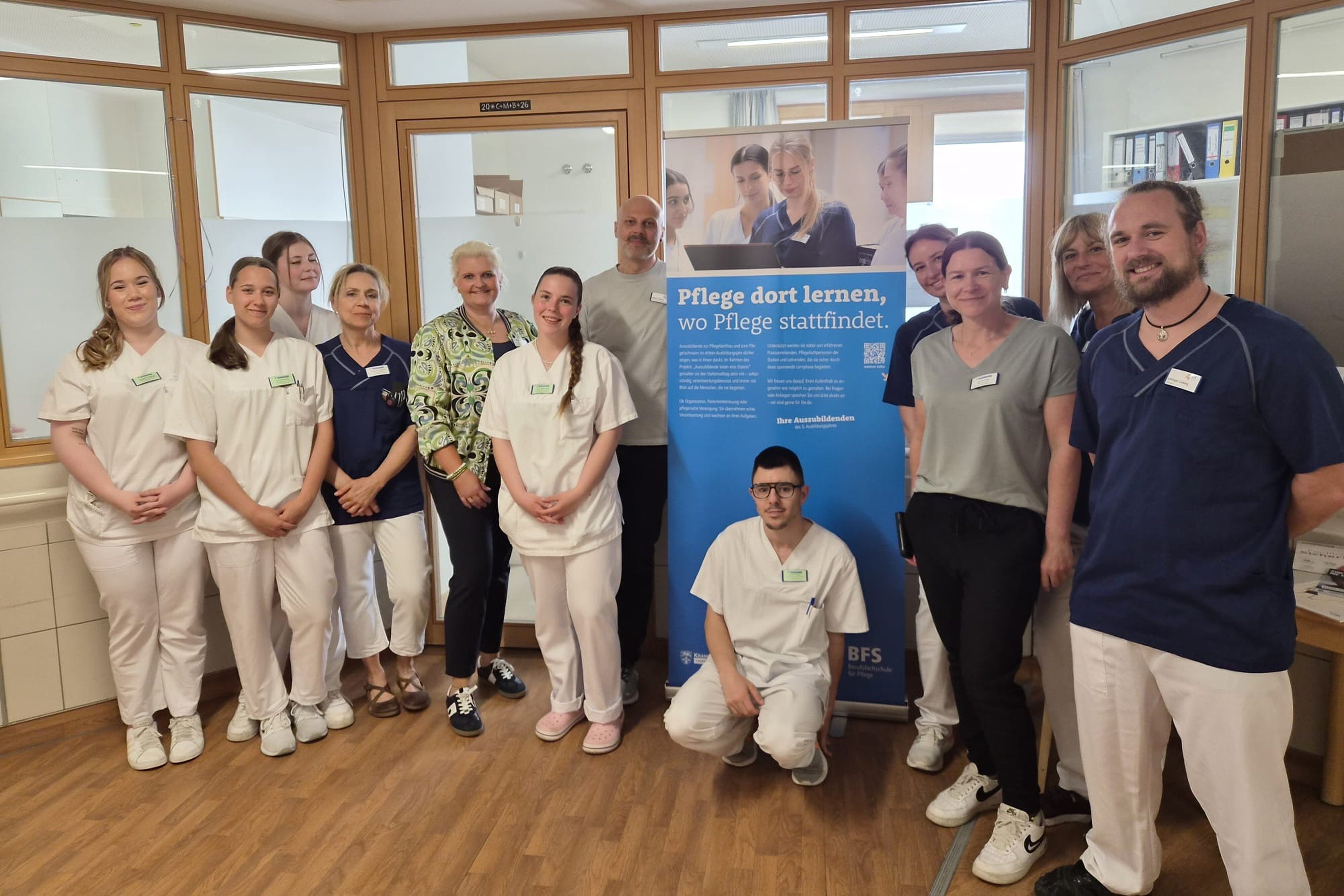 Azubis des dritten Lehrjahres im Gang der Station 2 in Weilheim vor Plakat mit dem Titel Auszubildende leiten eine Station.
